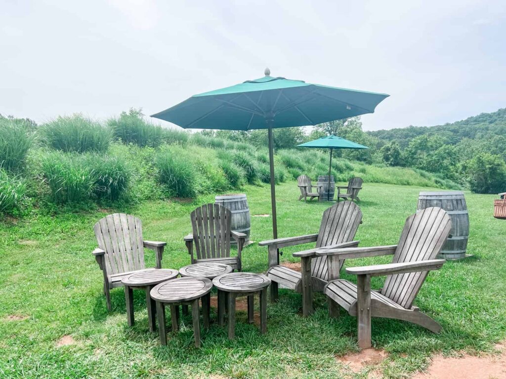 A cozy outdoor seating area with wooden chairs and tables under a green umbrella at Glen Manor Vineyards, inviting visitors to relax amidst the lush greenery of the surrounding hills.