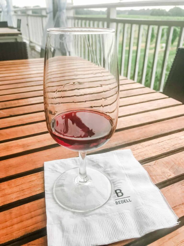 A glass of red wine resting on a wooden table, with a napkin featuring the Bedell logo underneath, overlooking a vineyard.