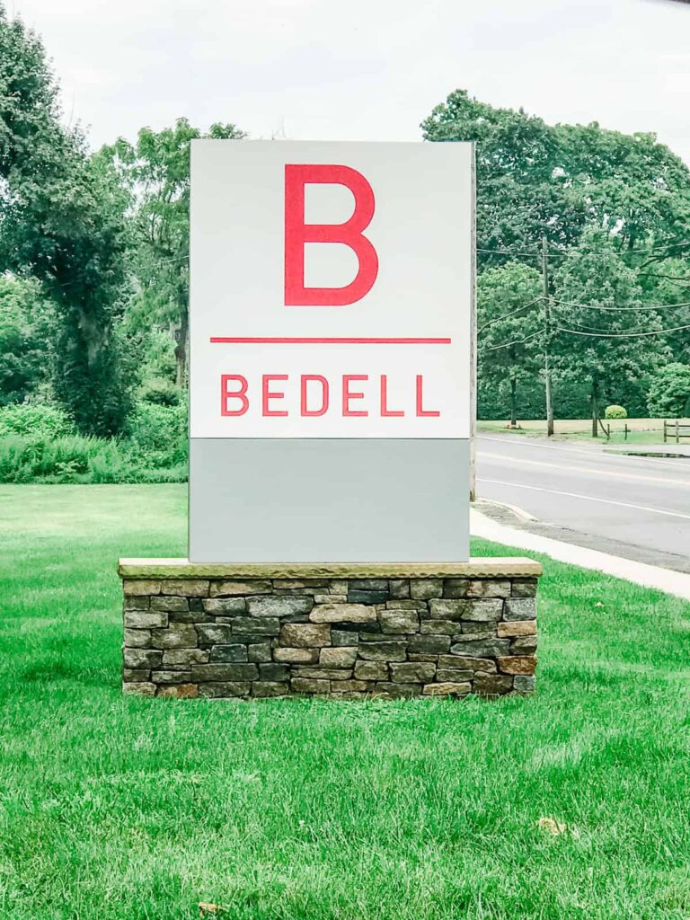 A large white and red sign with the letter "B" for Bedell Winery on a stone base, surrounded by green grass.