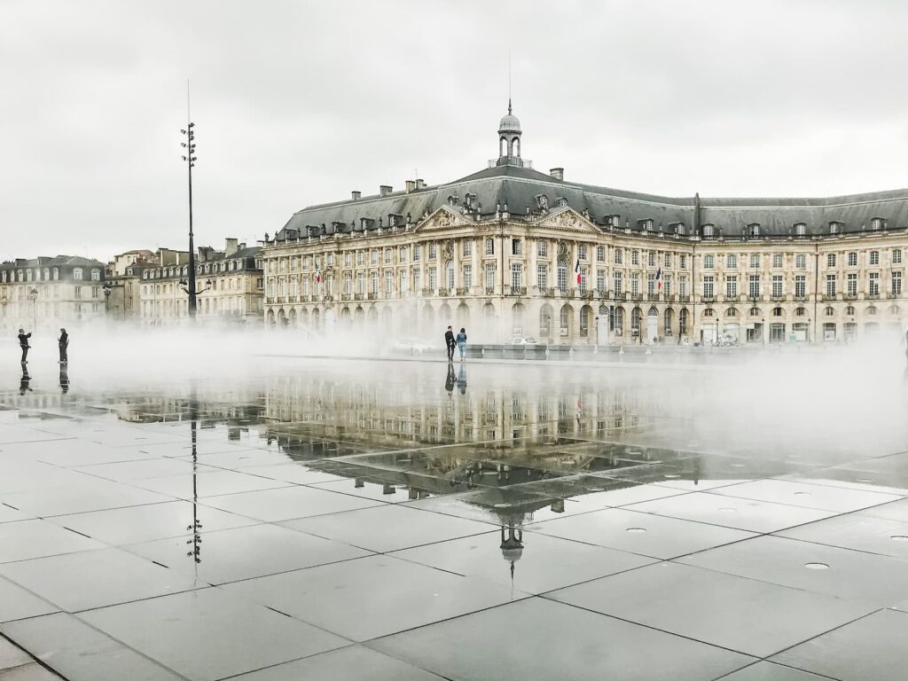 A grand classical building in Bordeaux, France, is reflected in the Miroir d'eau, a large water mirror that creates a dramatic scene with mist floating above its surface. On a cloudy day, a few people walk across the reflective plaza, adding scale to the architectural elegance.