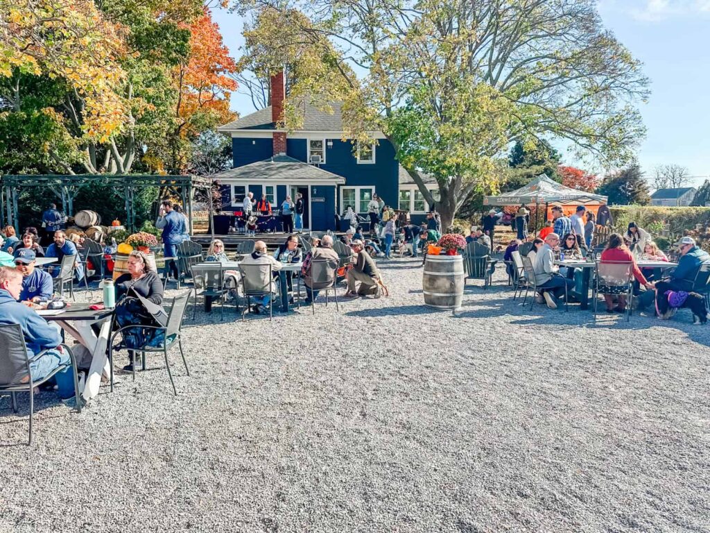 A lively outdoor seating area at Lenz Winery, where guests are enjoying food and drinks at scattered tables on a gravel patio. The setting includes a rustic blue house in the background, surrounded by vibrant autumn trees.