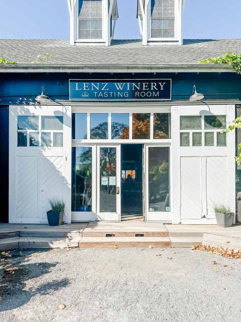 The front view of Lenz Winery's tasting room entrance, featuring large, white double doors with the "Lenz Winery Tasting Room" sign above, framed by greenery.