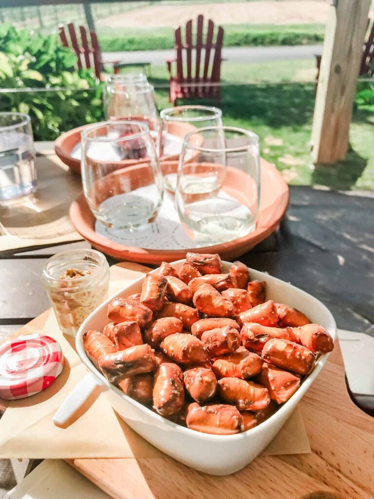 A white bowl filled with pretzel bites and a small jar of mustard on a wooden board, with a flight of wine glasses and Adirondack chairs visible in the background.