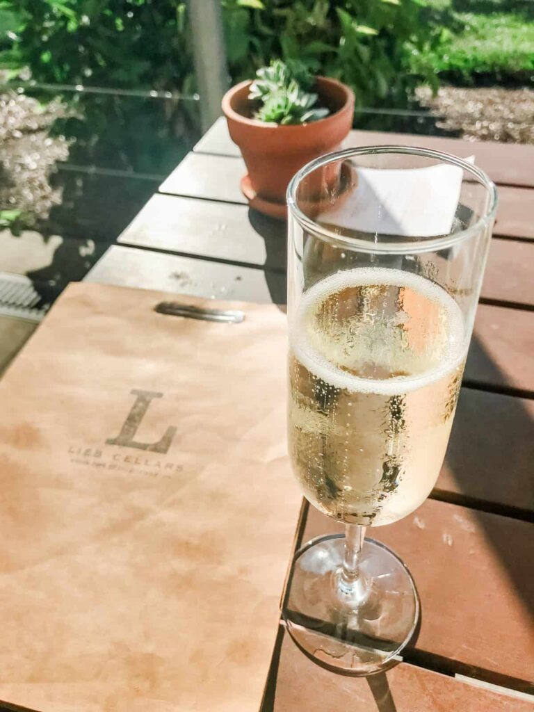 A flute of sparkling wine sitting on a wooden table, next to a tan menu folder labeled "Lieb Cellars," with a small plant in a pot visible in the background.