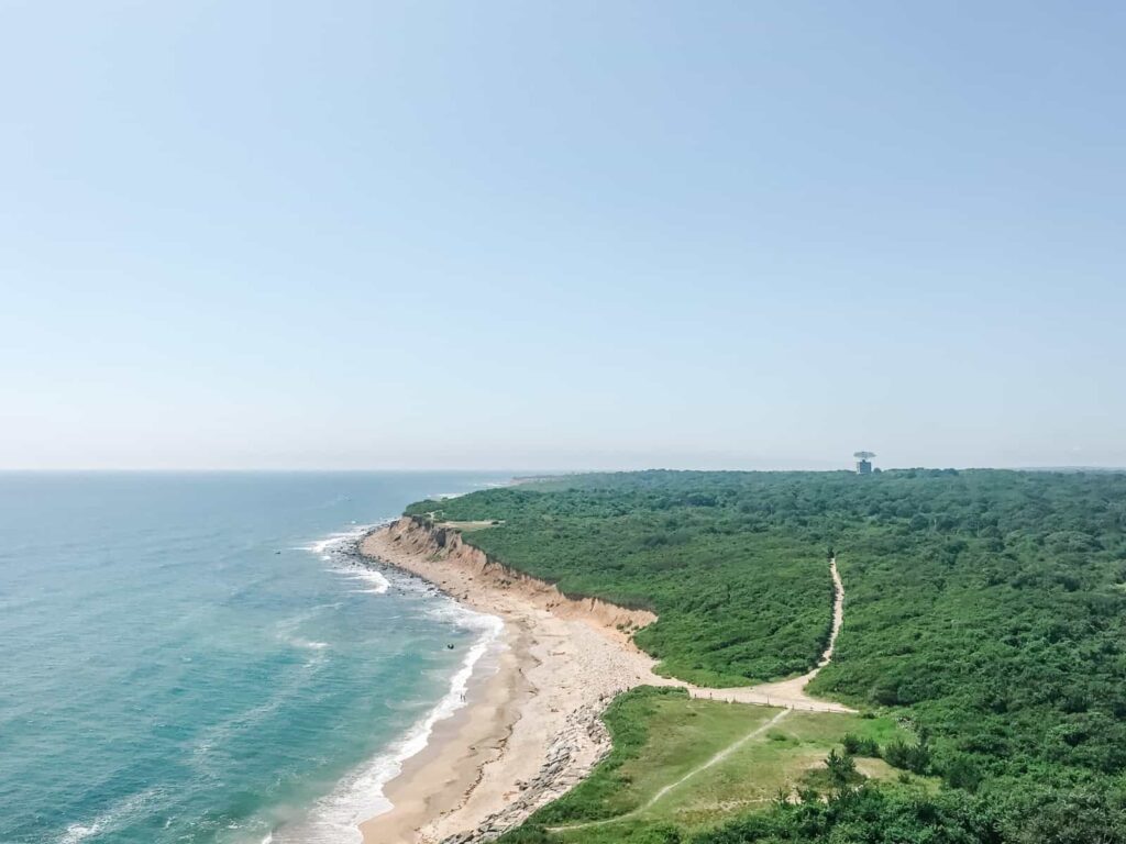 A coastal landscape on Long Island featuring a winding beach bordered by cliffs on the left and lush green foliage extending to the right under a clear blue sky. In the distance, a water tower is visible above the treeline.