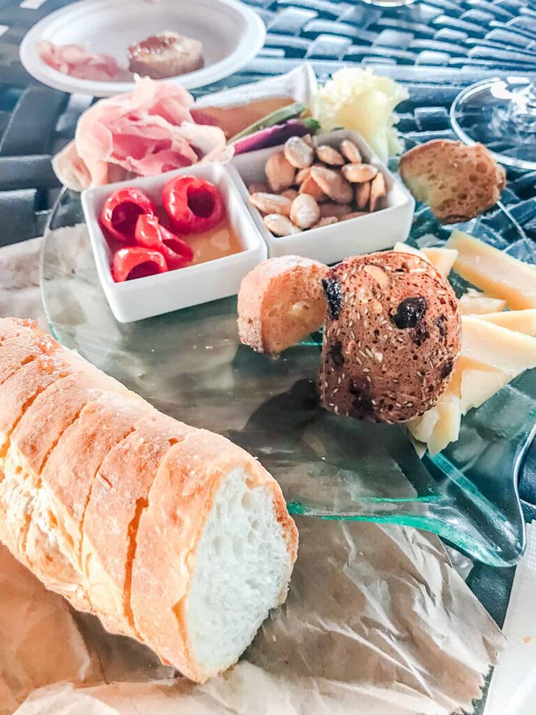A rustic charcuterie spread with sliced baguette, roasted nuts, prosciutto, mini red peppers, and a variety of cheeses on a glass platter, set on an outdoor table at Macari Vineyards.
