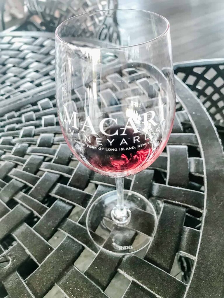 A partially filled glass of red wine with "Macari Vineyards, North Fork of Long Island, New York" printed on the glass, set on a black wrought iron outdoor table.