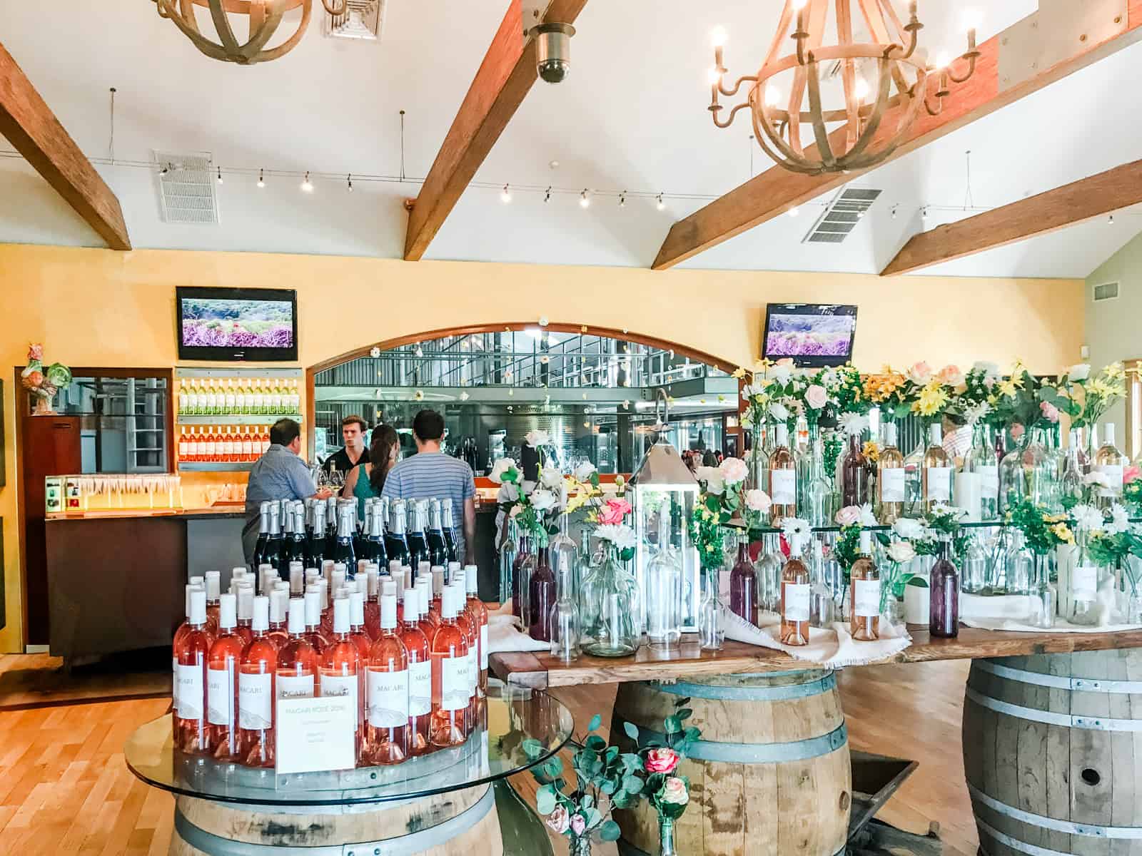 Inside the Macari Vineyards wine shop, a colorful display of rosé wine bottles, fresh flowers, and wine barrels, with a group of people enjoying a wine tasting at the counter in the background.