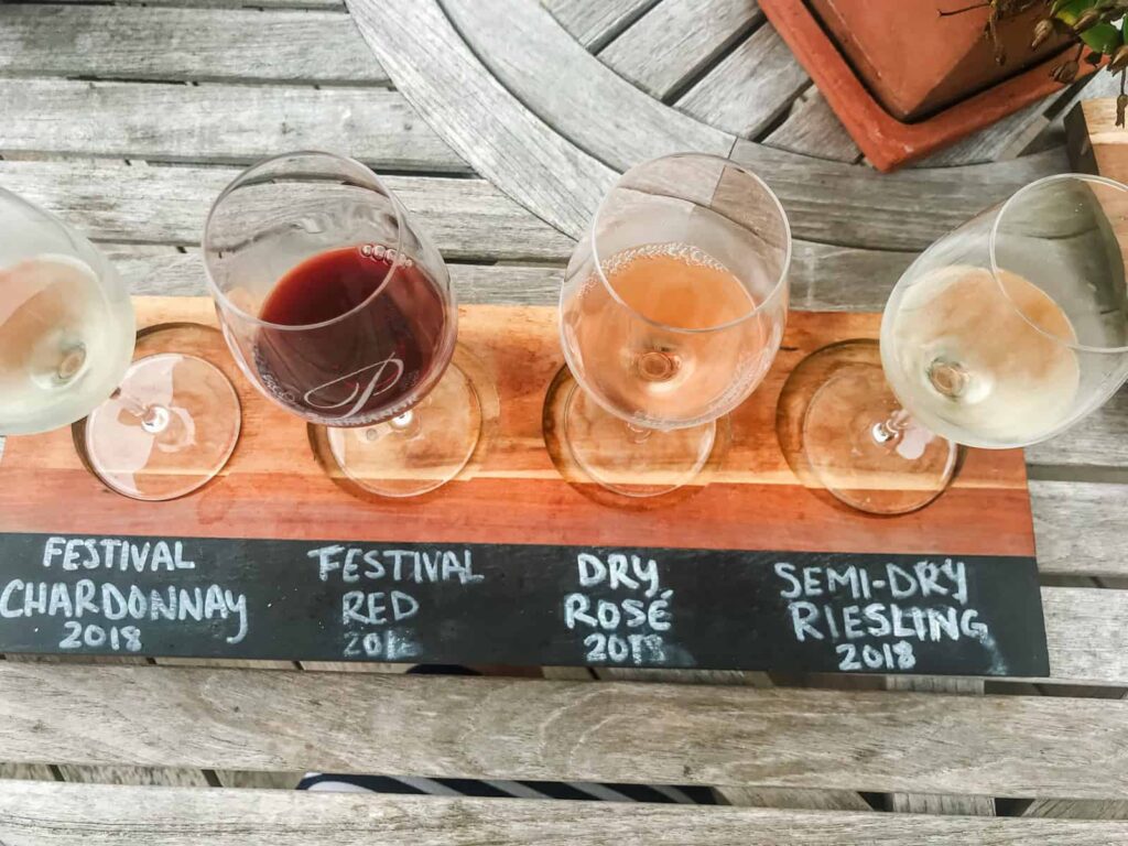 A wooden tray holding four glasses of wine, each labeled in chalk on the tray. The flight is placed on a rustic wooden table outdoors with a flower pot nearby.