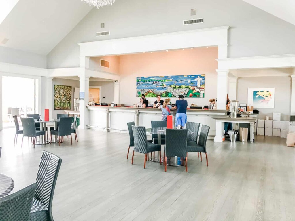A spacious, modern tasting room with high ceilings, white walls, and wooden furniture. There are several tables with black chairs and a bar counter at the back, where a few people are standing.