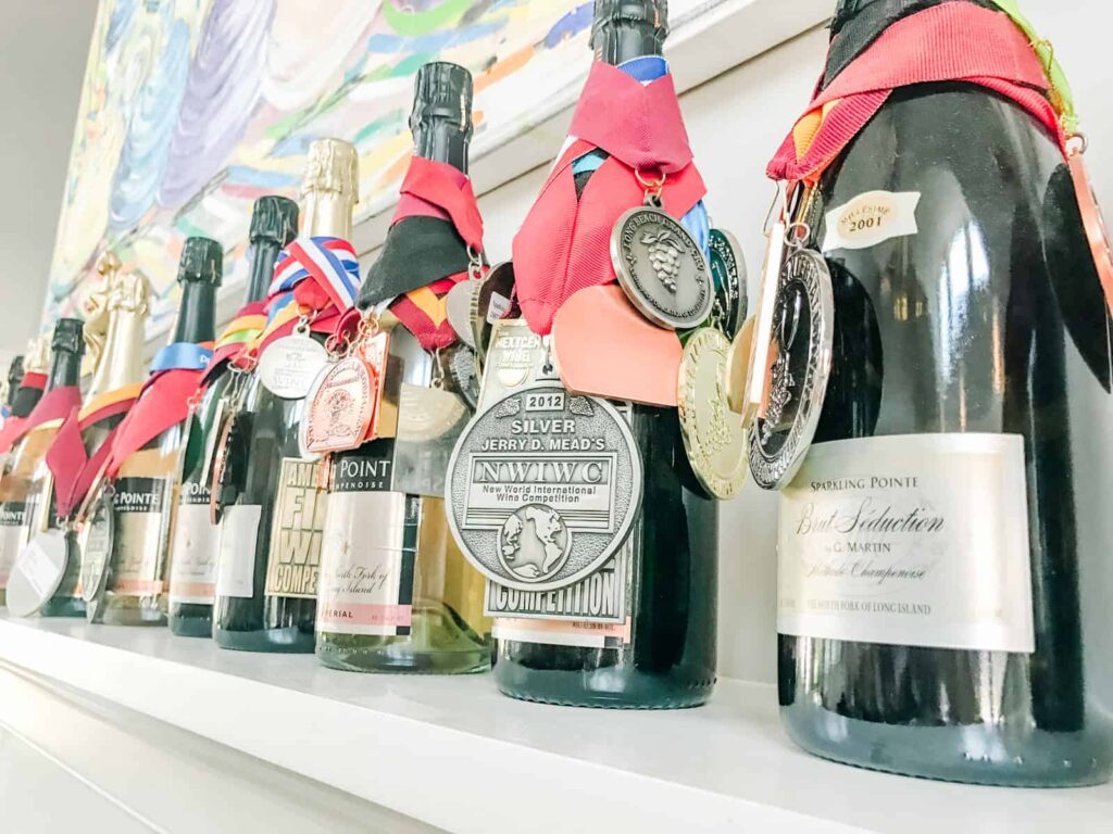 A close-up of Sparkling Pointe wine bottles lined up on a shelf, each bottle adorned with colorful ribbons and medals showcasing various awards won.