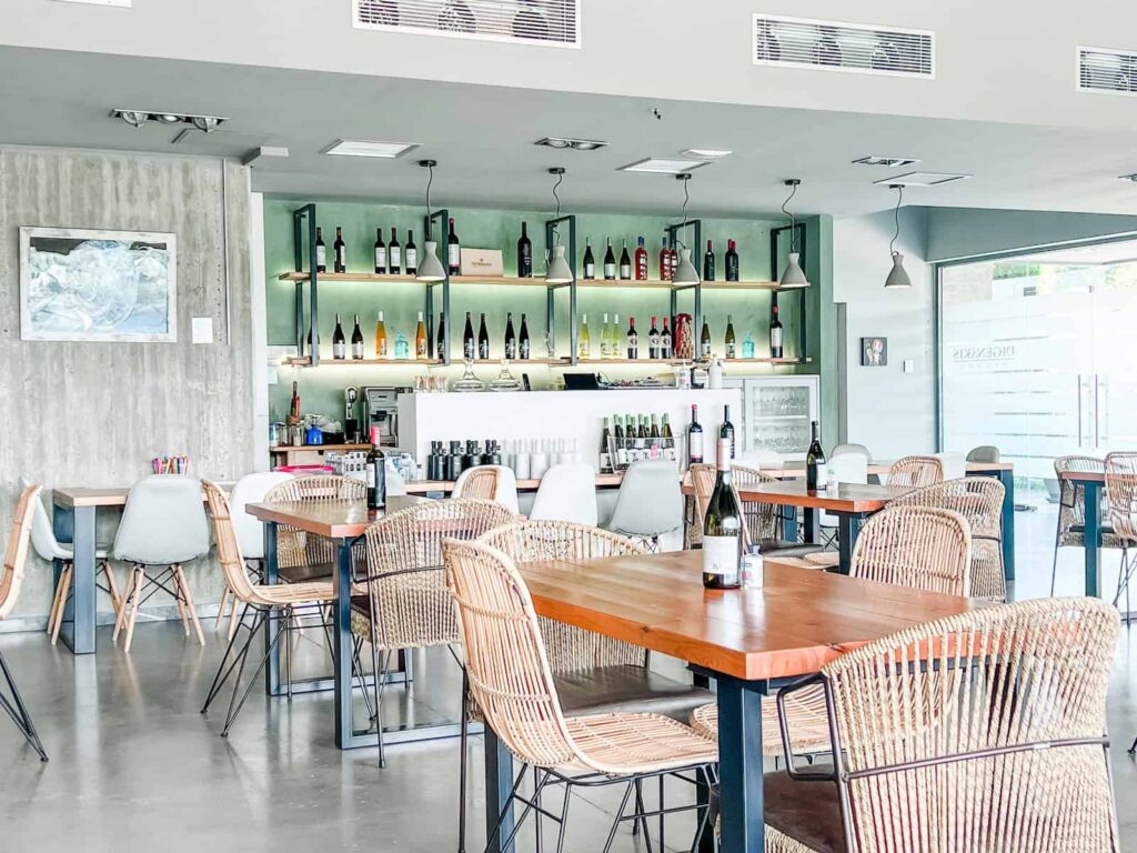 The contemporary interior of Digenakis Winery in Heraklion, Crete, showcasing a selection of wines on the bar shelf, wooden tables, and wicker chairs, all under the soft, natural light filtering through large windows.