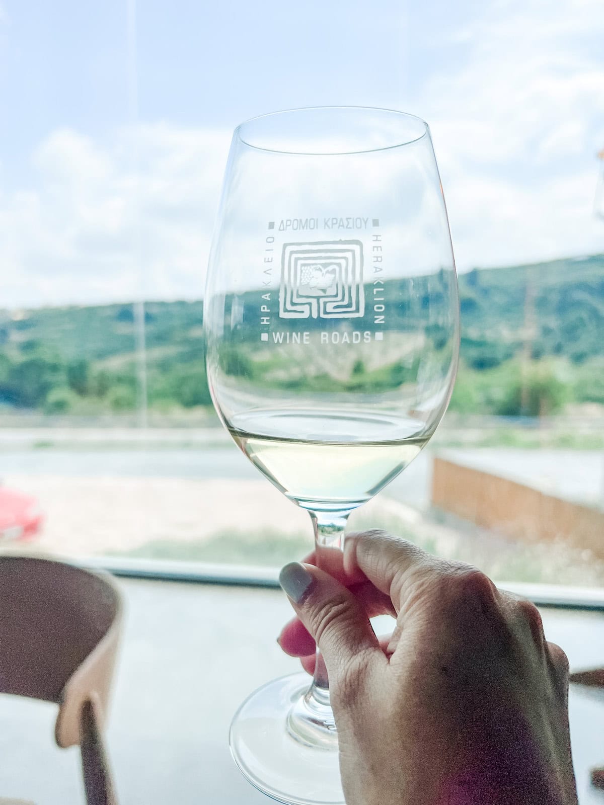 Close-up of a hand holding a clear wine glass engraved with the logo 'Wine Roads Heraklion' with a backdrop of the serene, green landscape of Crete seen through a sunlit window at Digenakis Winery.