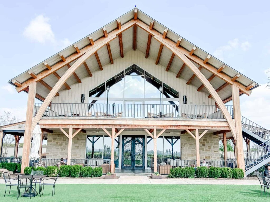 Modern winery building with a grand wooden A-frame structure, large glass windows, and an upper balcony overlooking a well-manicured lawn with patio seating available. The architecture blends rustic charm with contemporary design.