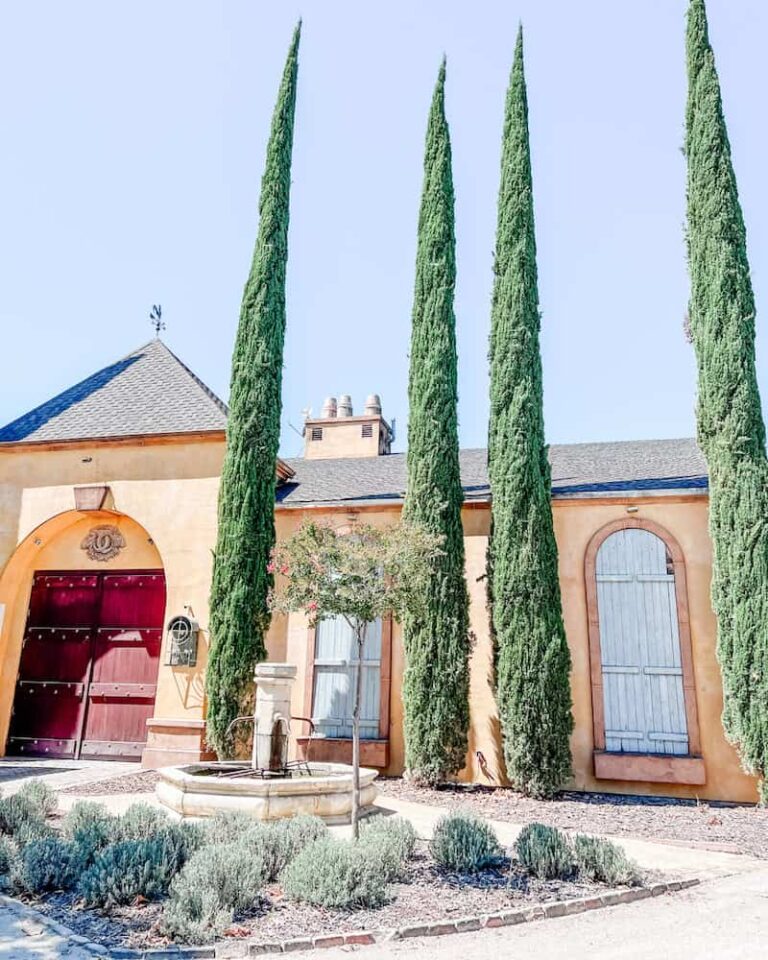 Facade of Demetria Estate winery with yellow stucco and red doors, accented by tall, slender cypress trees and a vintage water fountain in the foreground, invoking a sunny, Mediterranean vibe.