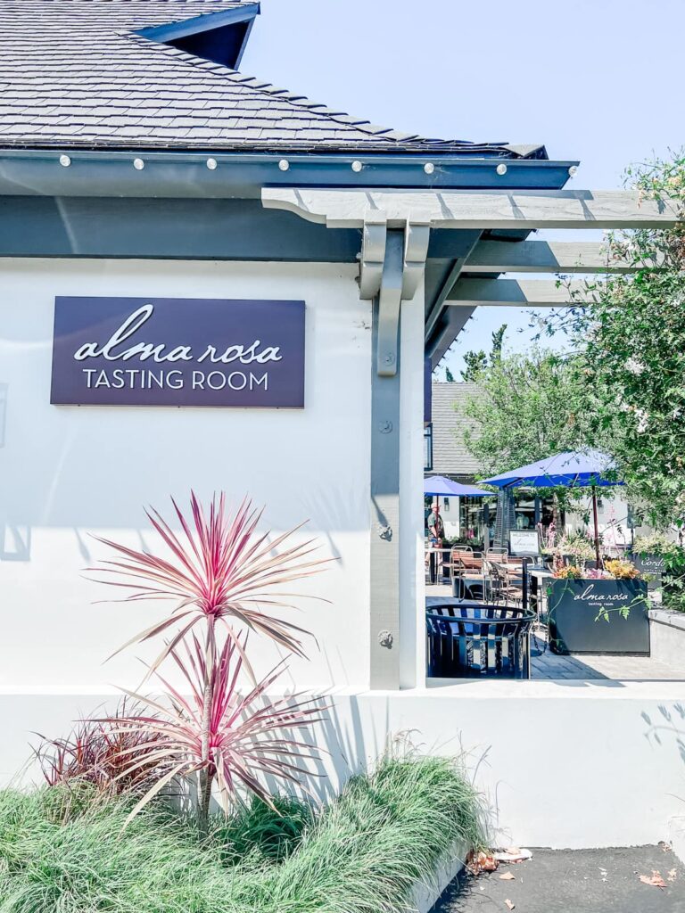 Signage of Alma Rosa Tasting Room mounted on a white wall with a vibrant pink plant in the foreground.