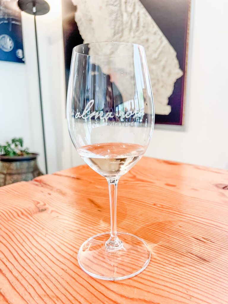 A close-up of a stemmed wine glass with the Alma Rosa logo, filled with white wine on a wooden table.