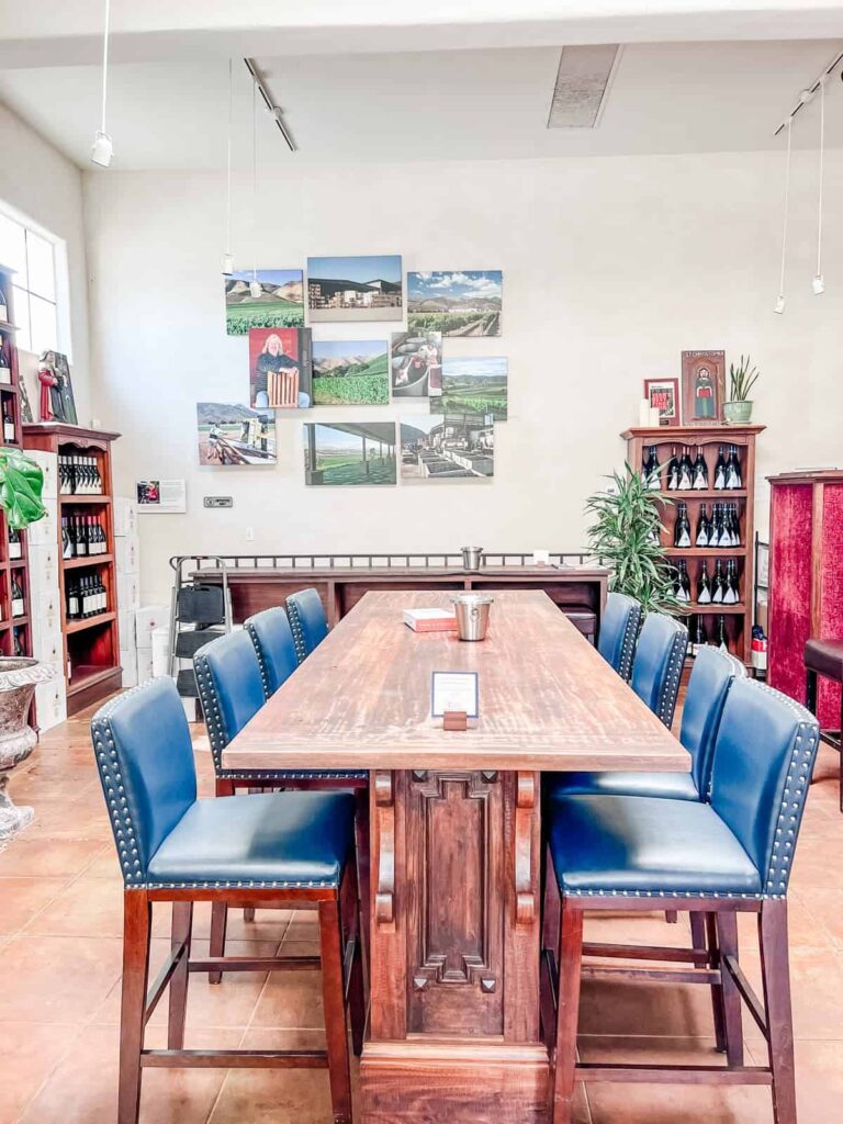 A spacious tasting room at Au Bon Climat featuring a large wooden table with blue leather chairs, surrounded by shelves of wine bottles and a wall collage of vineyard images.