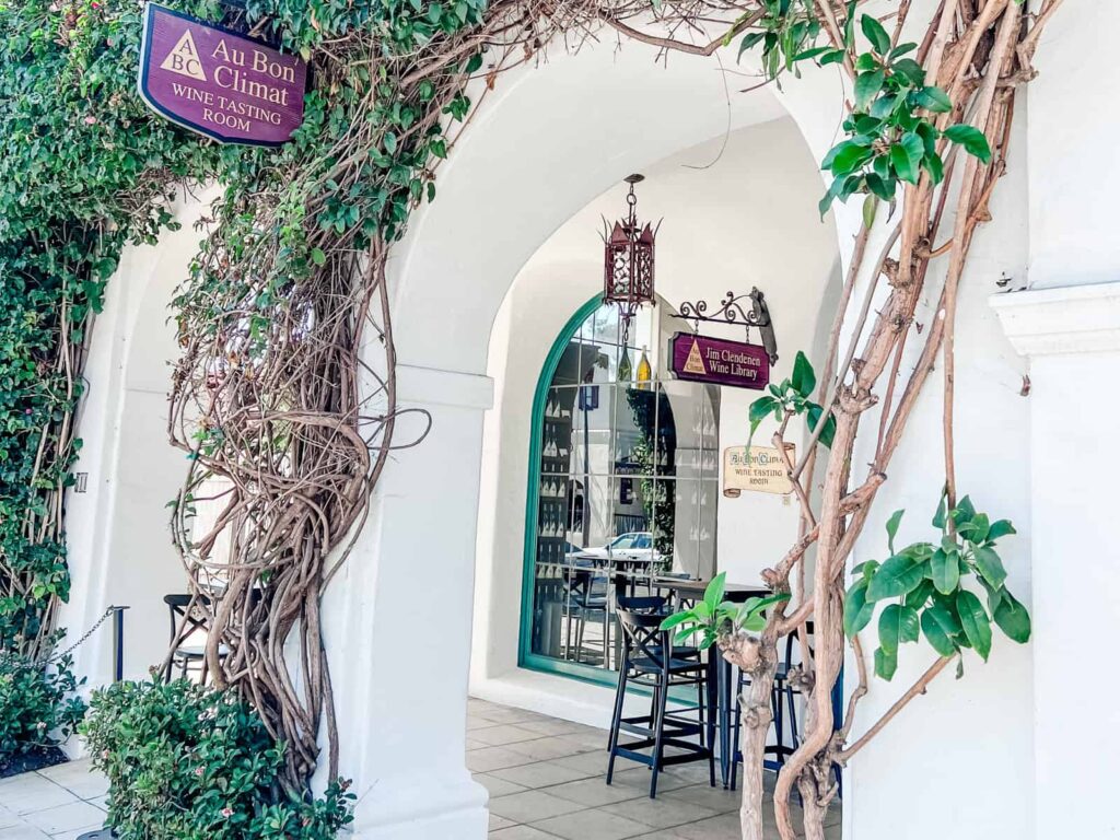 The vine-covered entrance of Au Bon Climat winery with a hanging sign for the wine tasting room and the Jim Clendenen wine library, inviting visitors into the arched doorway.