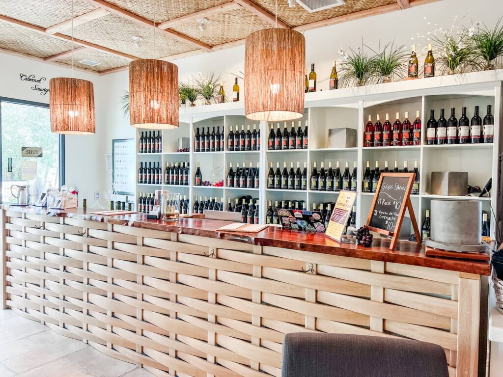Interior of Dascomb Cellars tasting room featuring a wooden bar, wine bottles on shelves, and hanging basket lights.