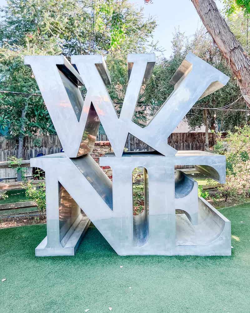 A large, metallic, three-dimensional sculpture of the word "WINE" with the 'W' and 'I' positioned higher than the 'N' and 'E', set on a green artificial turf at Malibu Wine.