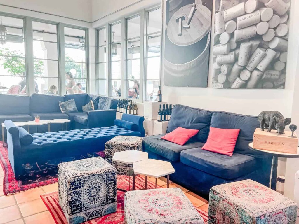 A cozy lounge area inside the Margerum Wine Company tasting room, with plush navy blue sofas and colorful ottomans, accentuated by wine-themed decor and large windows inviting natural light.