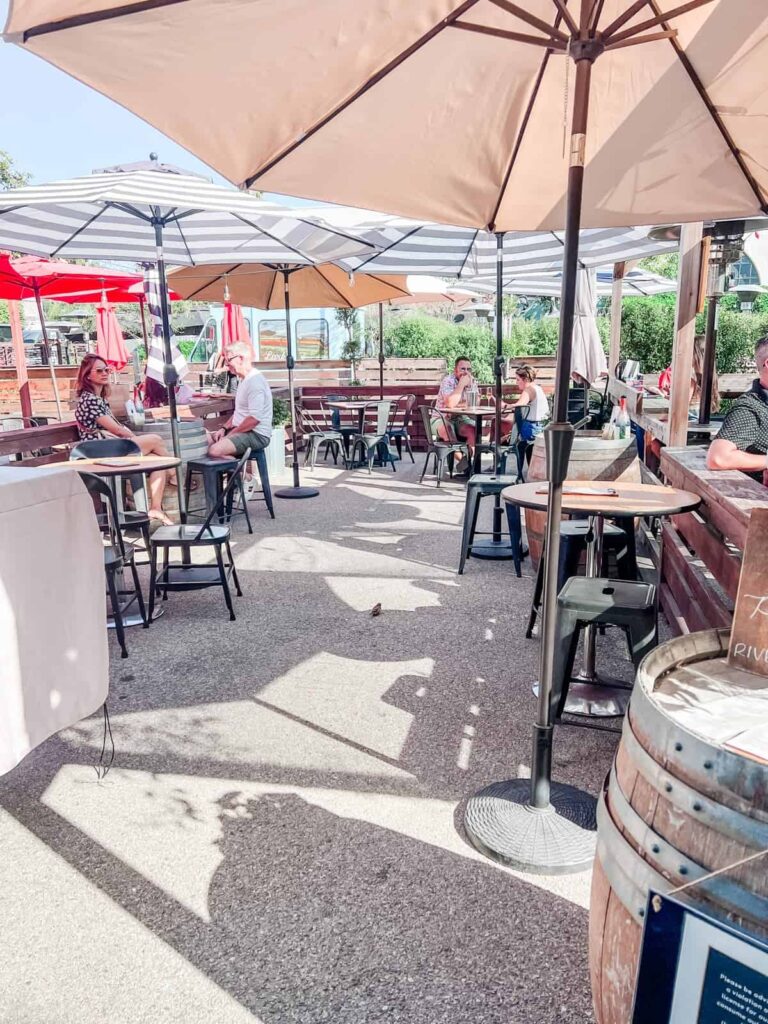 Patrons enjoying wine under the shade of beige umbrellas on a sunlit patio with wooden tables at Riverbench Winery, creating a relaxed outdoor tasting experience.