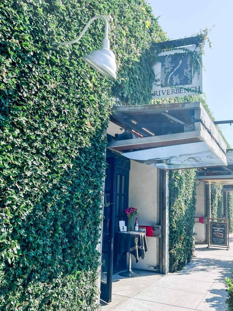 The ivy-covered entrance to Riverbench Winery's tasting room, marked by an elegant sign and outdoor seating, evoking a welcoming ambiance for visitors.