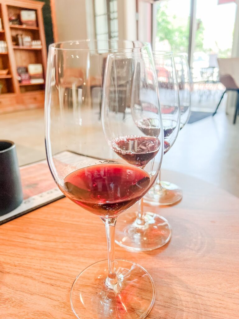 A trio of wine glasses at Toccata, with the closest showcasing a vibrant red wine, set on a wooden table in a bright tasting room.