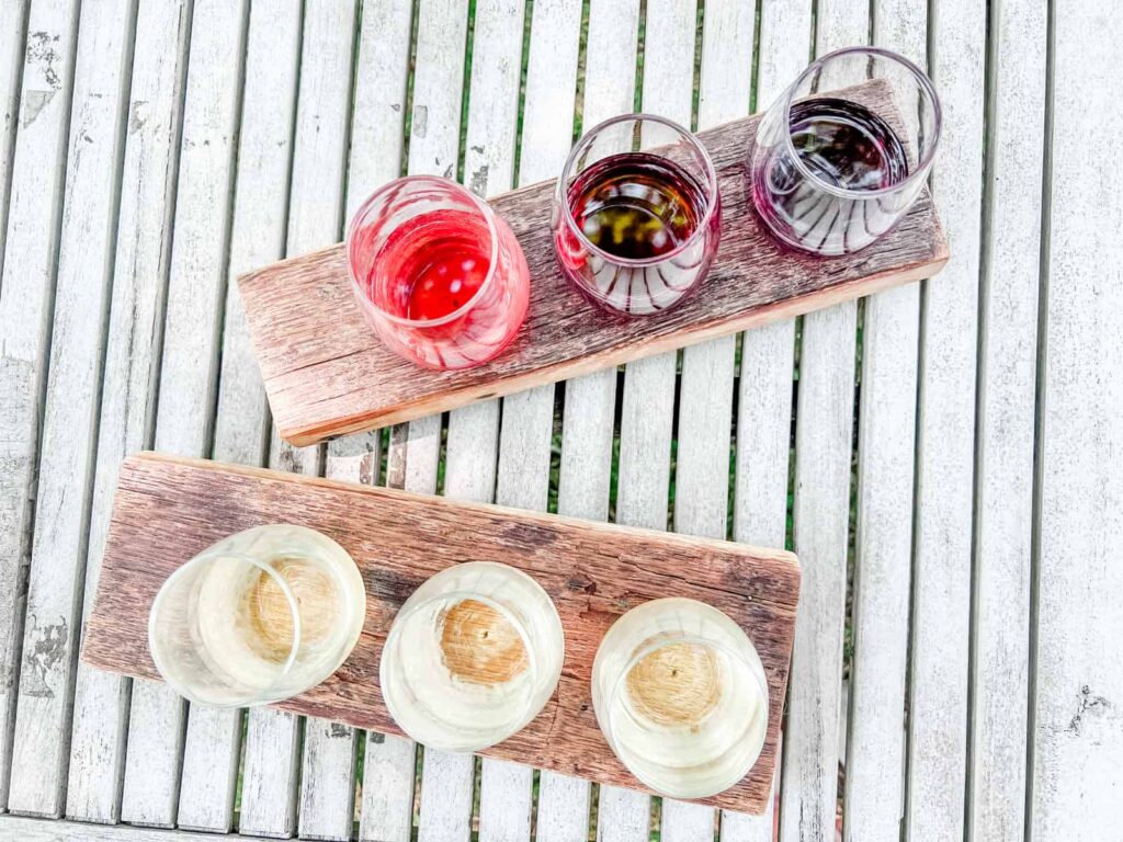 Two wooden flight boards with glasses of wine on a white painted wooden table: the top board holds a selection of red wines, and the bottom board presents white wines, ready for a tasting experience.