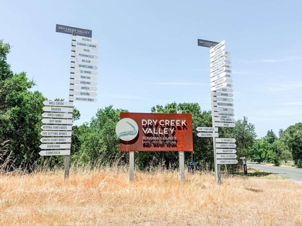 Large welcome sign for Dry Creek Valley AVA in Sonoma County, surrounded by tall directional posts listing local wineries like Ferrari-Carano, Mazzocco, Ridge, and Lambert Bridge.