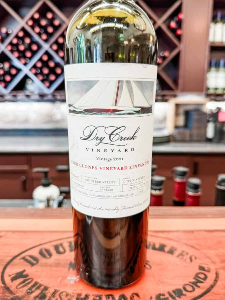 Close-up of a 2021 Dry Creek Vineyard Zinfandel bottle labeled "Four Clones Vineyard," with wine racks and bottles in the blurred background.