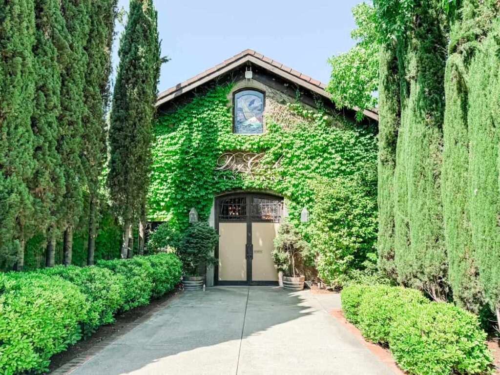 Ivy-covered entrance to Dry Creek Vineyard framed by tall trees and manicured hedges, with a sign above the door and a serene, inviting pathway.
