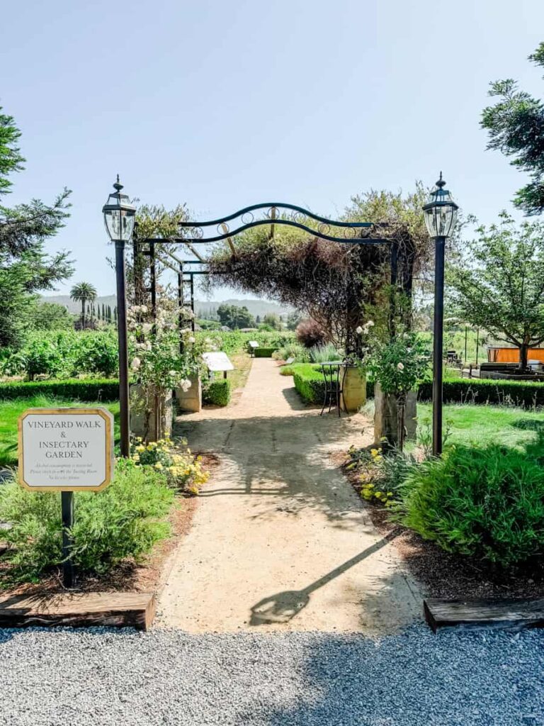 Decorative iron archway labeled "Vineyard Walk & Insectary Garden" leading into a landscaped garden with paths and vineyard views in the distance.