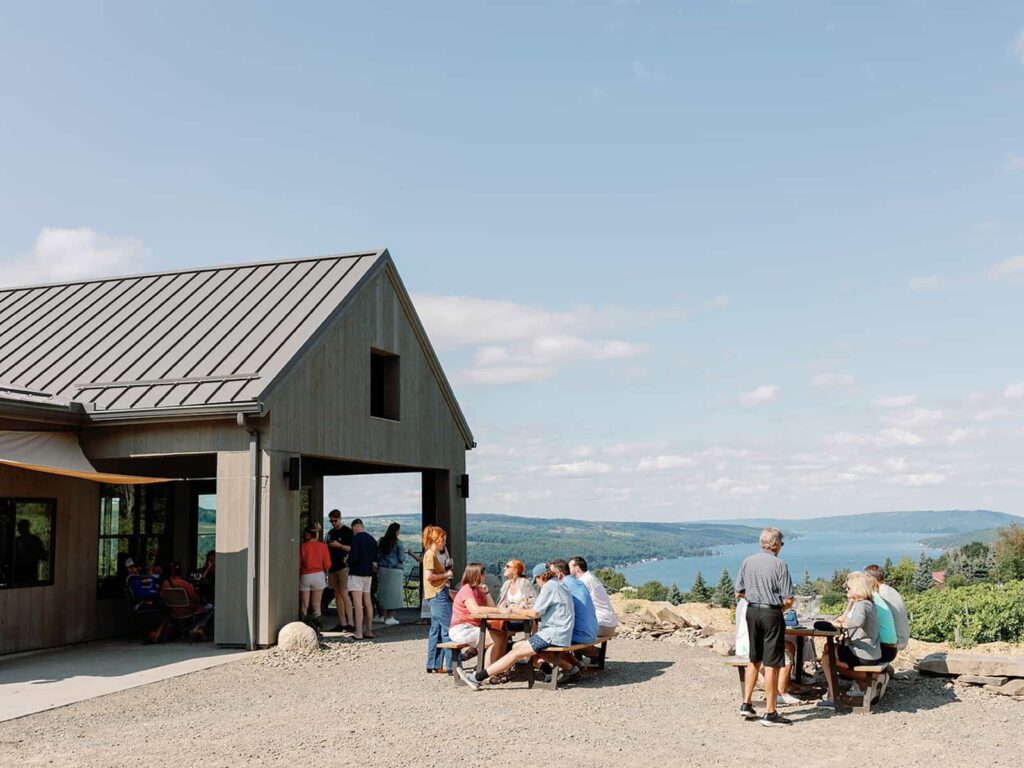 A scenic view of a winery patio at Living Roots on Keuka Lake, with groups of people seated at tables enjoying wine under a clear blue sky. The lake and surrounding hills are visible in the background.