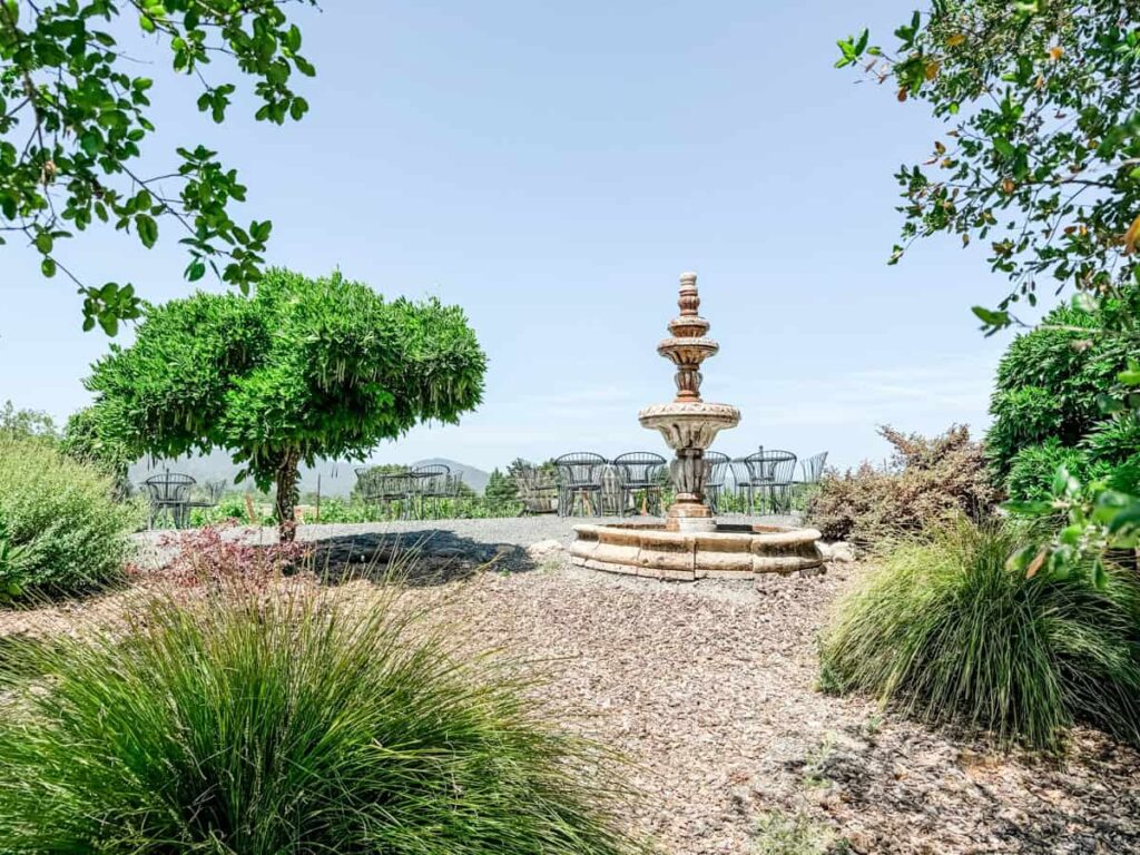 Stone fountain centerpiece in a tranquil garden area at Mazzocco Winery, with bistro tables overlooking the vineyard and distant hills.