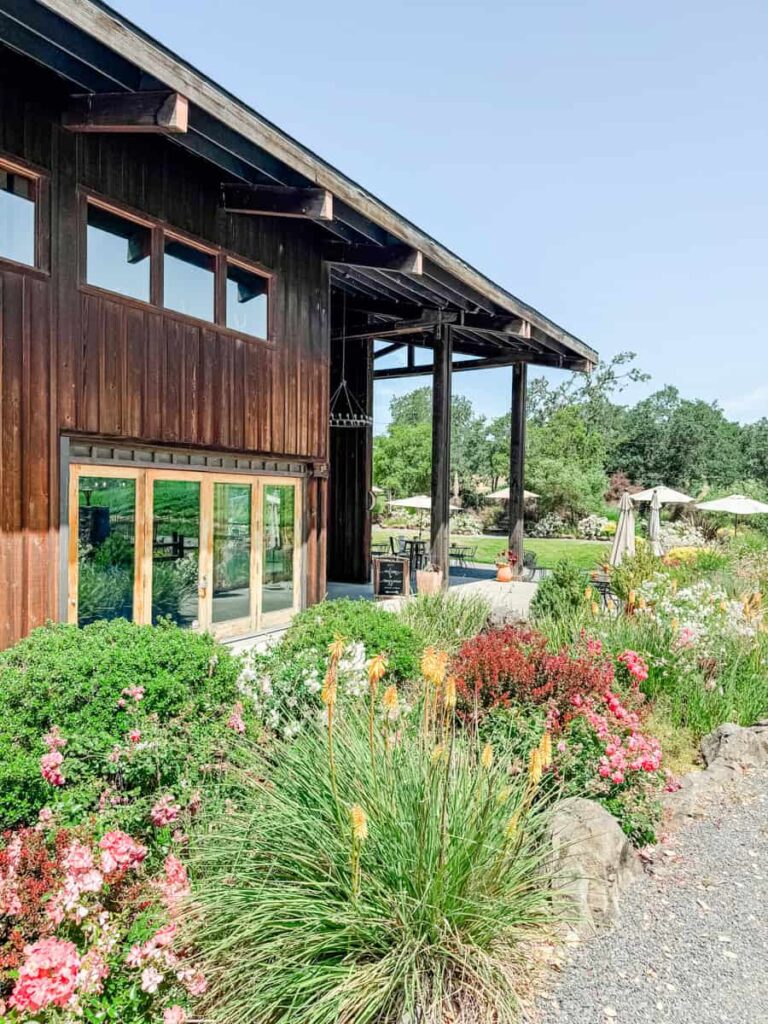 Colorful landscaping and flowers lining the exterior of Mazzocco Winery’s wooden tasting room, with large windows and a gravel path.