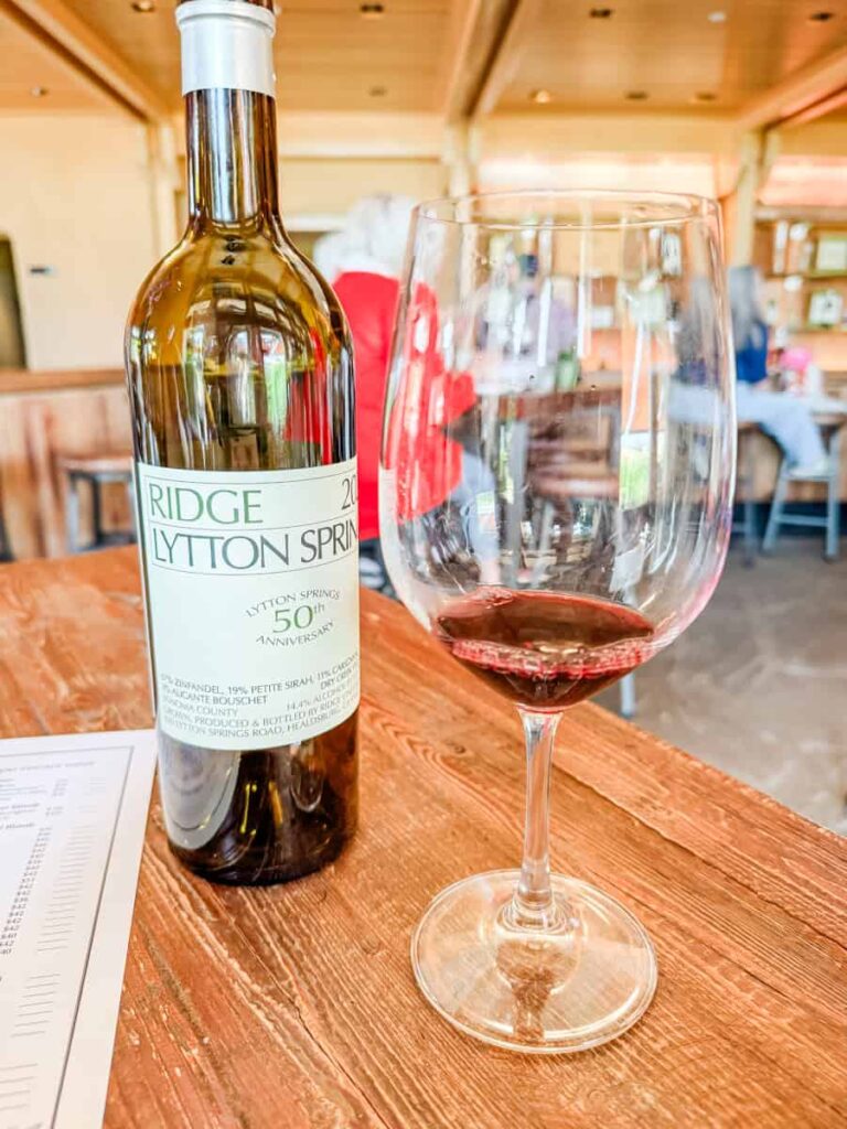 Bottle of Ridge Vineyards Lytton Springs 50th Anniversary wine next to a glass of red, sitting on a rustic wooden tasting room table.
