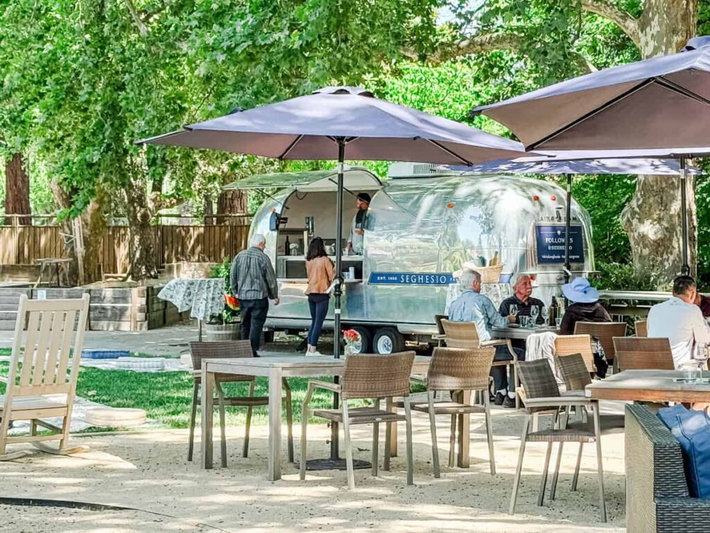 Guests enjoy wine tasting under large umbrellas in the shaded outdoor seating area of Seghesio Family Vineyards, with a shiny Airstream trailer wine bar in the background.