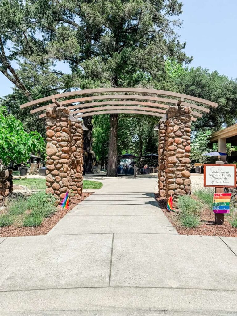 Stone archway entrance with a sign reading “Welcome to Seghesio Family Vineyards Tasting Room,” surrounded by trees and rainbow pride flags.