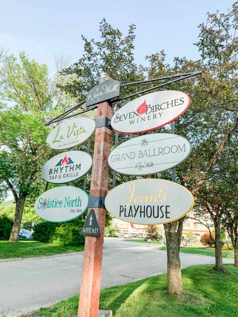 Wooden post with oval signs pointing to Seven Birches Winery, La Vista Italian restaurant, Jean’s Playhouse, and other amenities at RiverWalk Resort in Lincoln, New Hampshire.