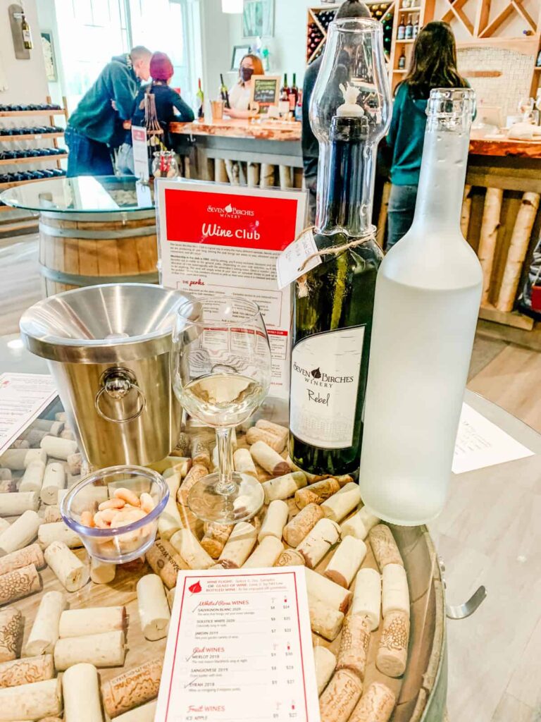 A wine tasting setup at Seven Birches Winery featuring a glass of white wine, a bottle of Rebel red wine, and a wine club information card, surrounded by a decorative arrangement of wine corks and a bowl of peanuts on a glass table.
