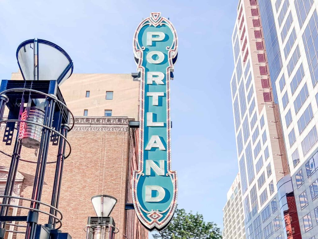 A vertical, turquoise-colored marquee sign with the word "PORTLAND" in large white letters, set against a backdrop of buildings in downtown Portland, Oregon.