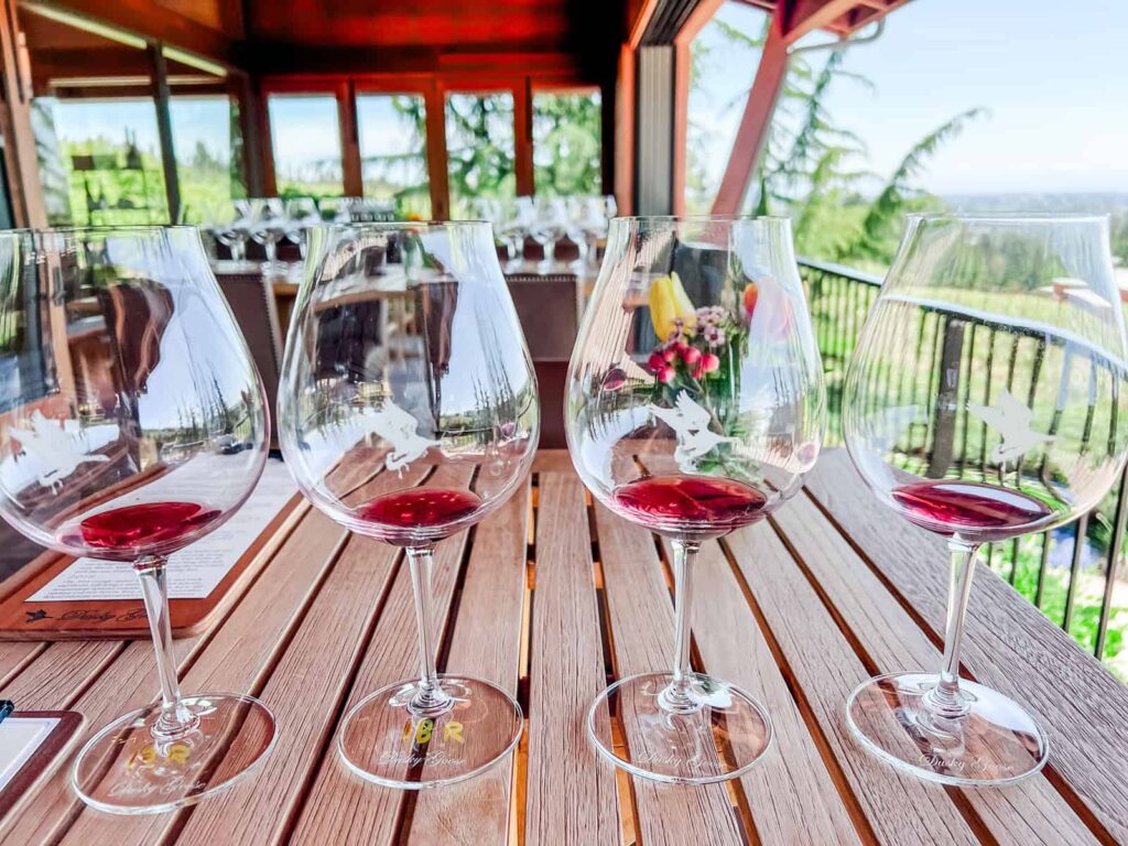 Four wine glasses with small amounts of red wine sitting on a wooden table in a tasting room overlooking the scenic vineyards of the Willamette Valley, Oregon.