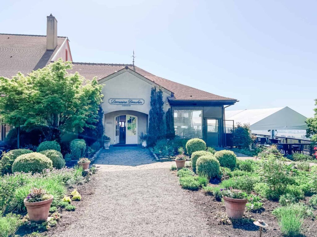 A classic winery building with light-colored walls, neatly landscaped gardens, and a gravel walkway leading to the entrance. The building is surrounded by vibrant greenery and planters.