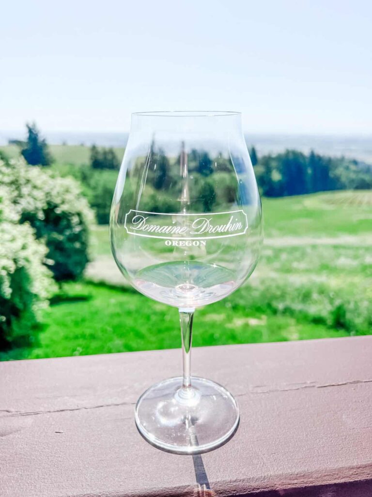 A clear wine glass with the "Domaine Drouhin Oregon" logo is held up against a backdrop of green vineyards and a bright, sunny day.
