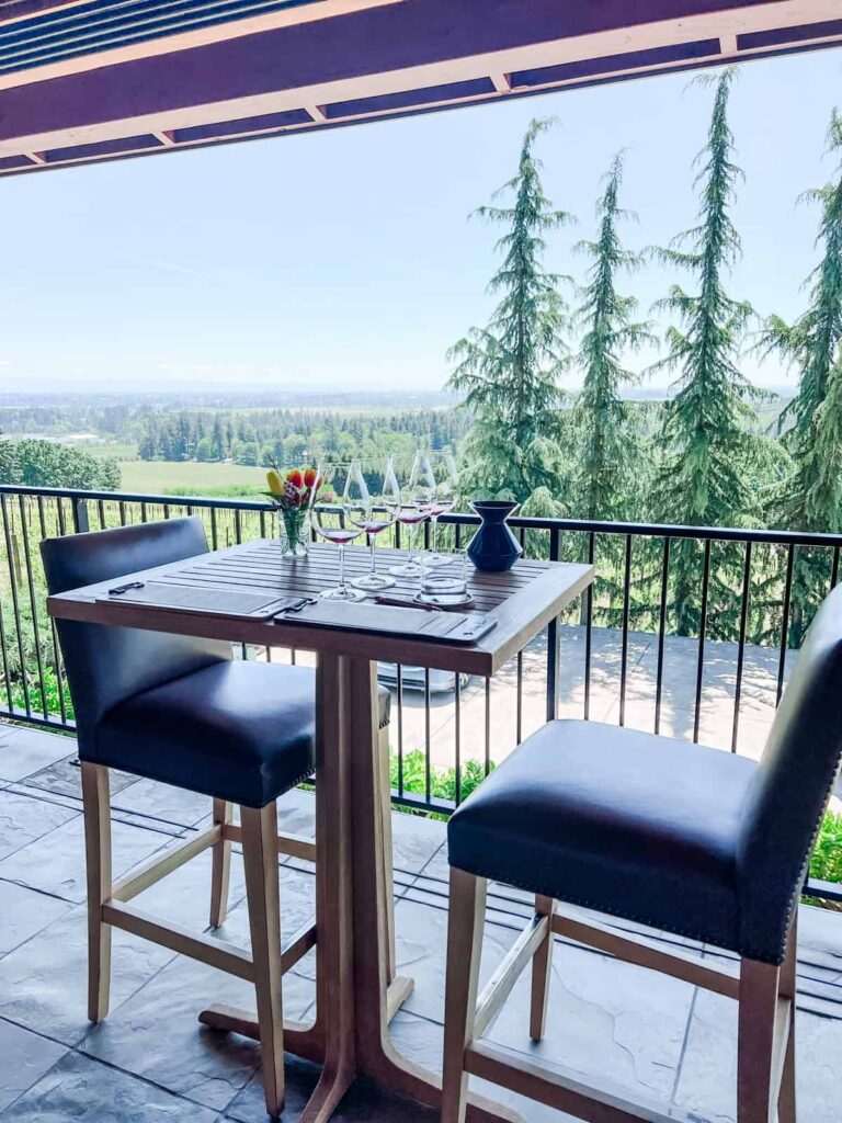 A high-top wooden table with two chairs on a covered patio, overlooking a view of pine trees and vineyards under a clear blue sky.