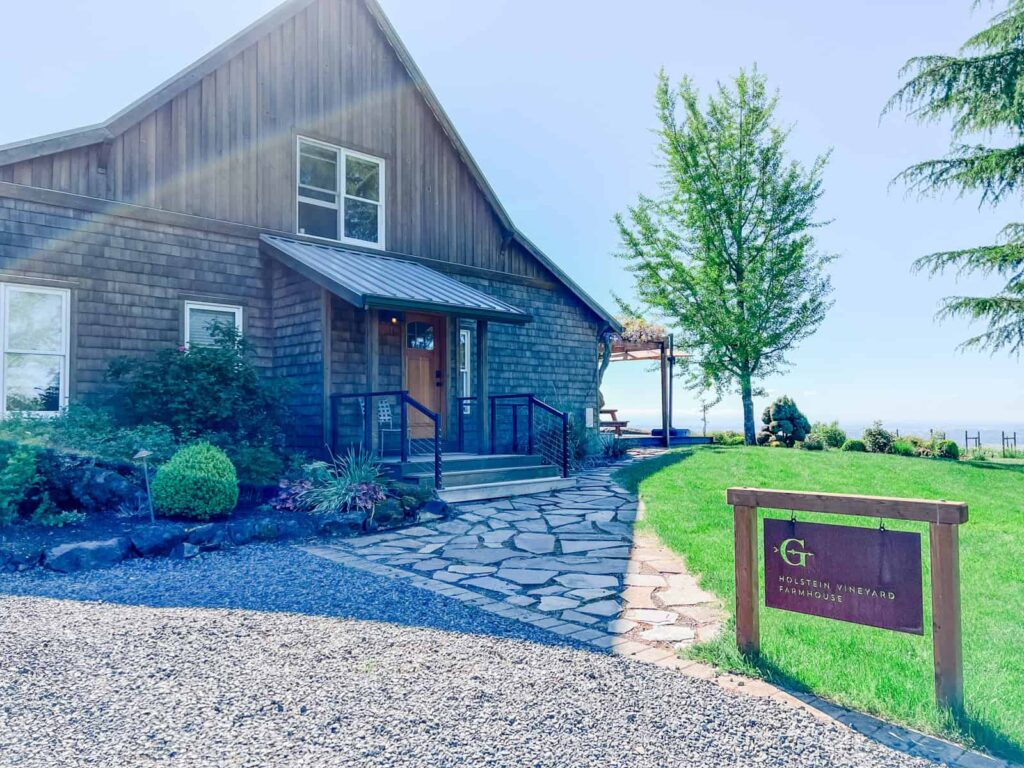 A rustic farmhouse-style building with stone walkways and a sign that reads "Holstein Vineyard Farmhouse," surrounded by neatly landscaped greenery.