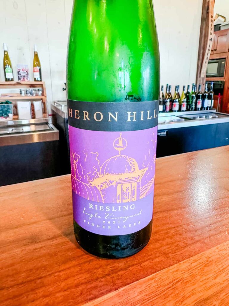 A bottle of Heron Hill 2021 Riesling from the Ingle Vineyard in the Finger Lakes, displayed on a wooden counter with a purple and black label showing an illustration of a dome structure.