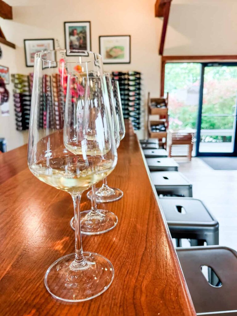 A row of three wine glasses with white wine sits on a polished wooden bar, with a backdrop of wine racks and framed pictures in the Heron Hill tasting room.